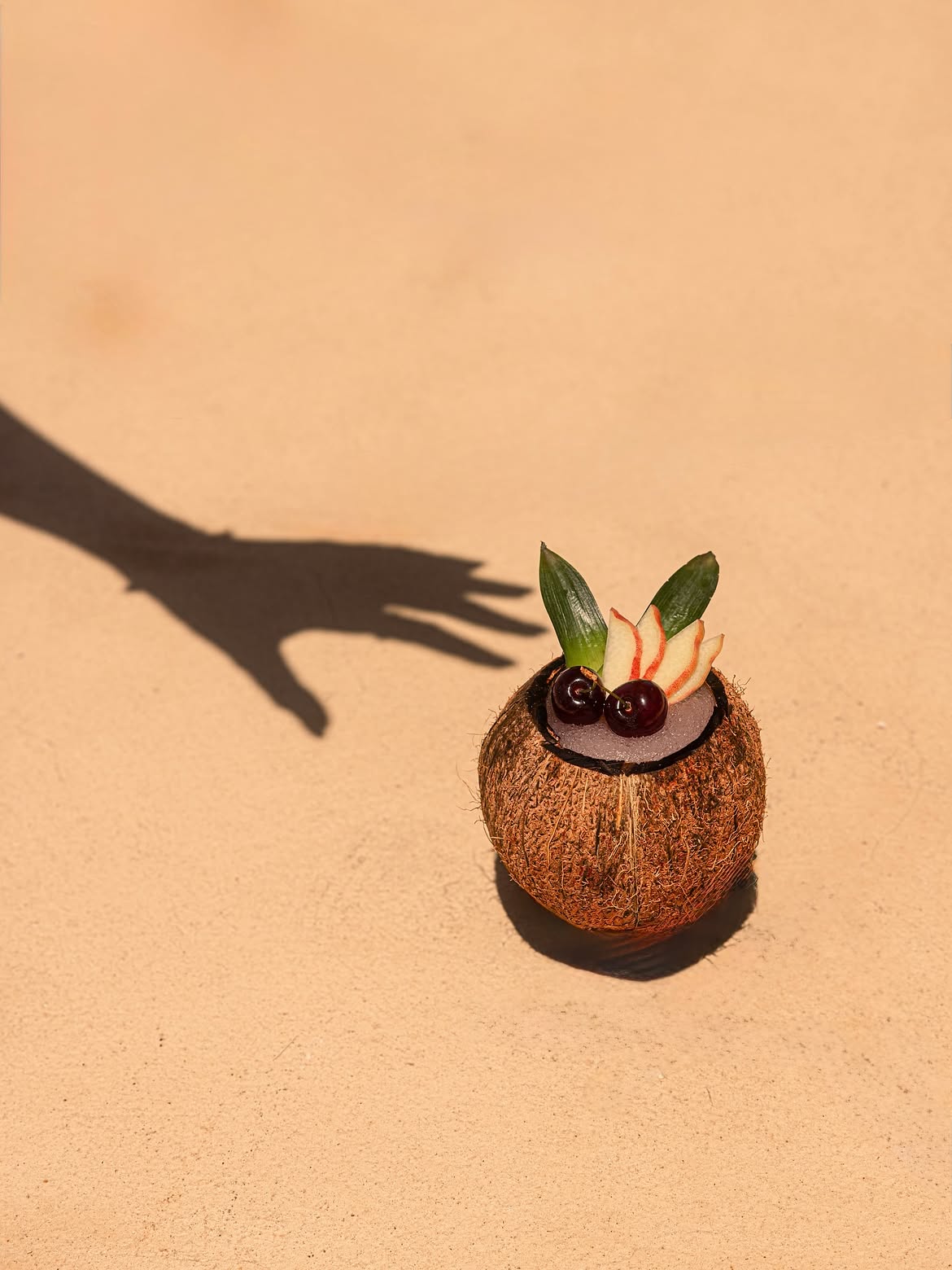 Coconut cocktail on warm sand with a hand's shadow reaching for it