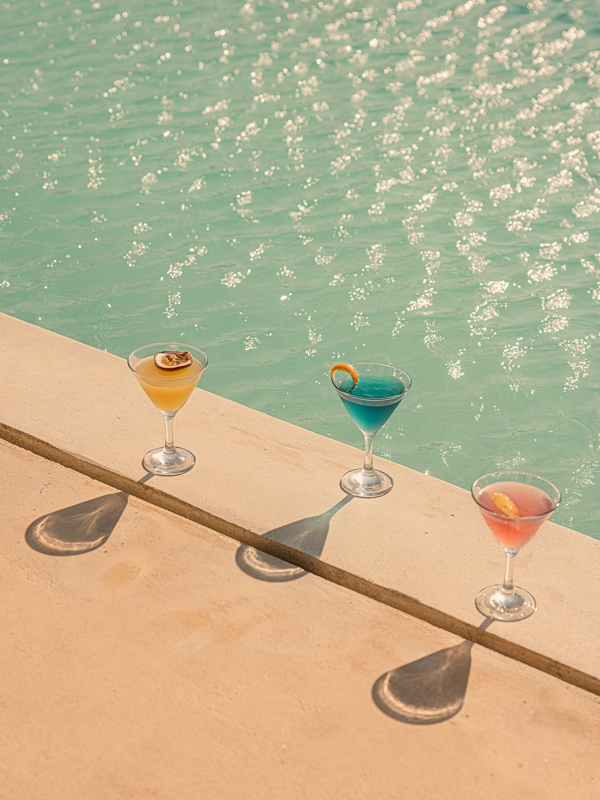 Three cocktails lined along the pool coping casting long shadows