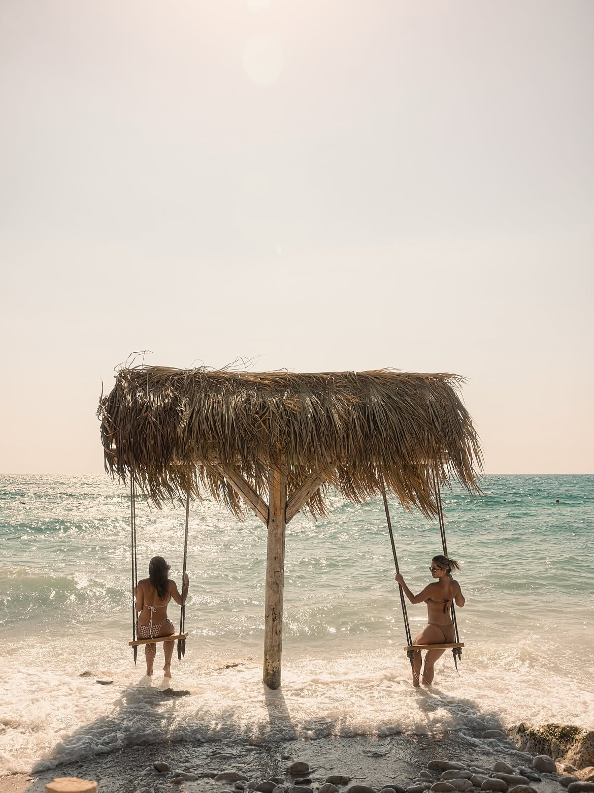 Two guests on rope swings under a thatched shade in the shallows