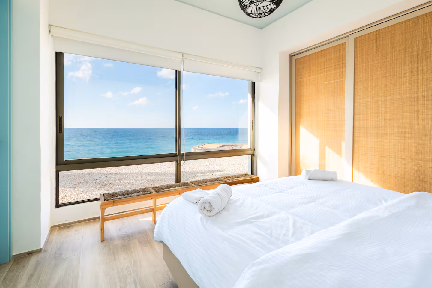 White chalet bedroom with a wide window onto the pebble beach and turquoise sea